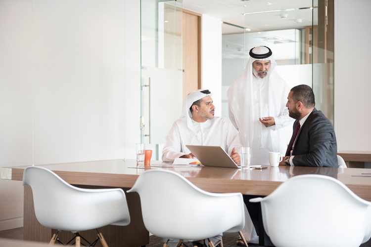 A photo of serious Middle Eastern businessmen discussing. Traditional Arab Emirati men in thobe talk to man in western suit. Middle East professional business people are in a meeting with laptop at conference table, Dubai, United Arab Emirates.